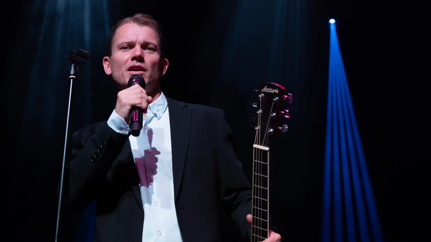 Mads synger i mikrofonen i blåt scenelys med vinplet på skjorten